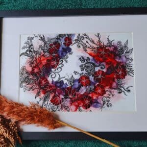 A framed artwork featuring a vibrant wreath of red and purple flowers with intricate black swirls and details on a white background. The frame is black with a white mat. Dry botanical elements and a logo reading "AewaCreations" are visible in the foreground.