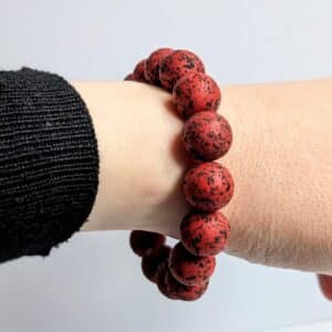 A person's wrist is shown wearing a red bead bracelet with black speckles. The person is dressed in a black, long-sleeve shirt, and the background is a plain, light-colored surface.